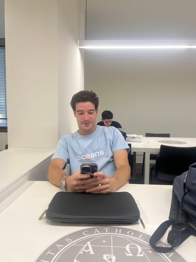 This image depicts an indoor scene within the Universidade Católica Portuguesa - Porto Campus, located in Foz do Douro, Aldoar, Foz do Douro e Nevogilde, Portugal.
In the foreground, a young adult male with dark brown hair and light skin is seated at a white table. He is wearing a light blue t-shirt with the word "oceans" and an orange horizontal line printed on the chest. On his left wrist is a silver wristwatch, and on his right wrist is a thin silver bracelet. He holds a dark-colored smartphone, equipped with a back grip accessory, in both hands, looking down at its screen with a slight smile. On the table in front of him, a black zippered case is visible, positioned next to a circular grey mat displaying the "UNIVERSITAS CATHOLICA" emblem. A dark-colored bag with a visible strap is partially present on the far right edge of the table.
In the mid-ground, another young adult male with dark hair is seated at a white table, positioned behind and to the right of the foreground subject. He wears a dark top and is looking down at a laptop. On his table, a white box and a stack of white rectangular items are present. Black office chairs with armrests are situated around this table.
The room features plain, light-colored walls and is illuminated by a prominent, linear LED light fixture mounted horizontally on the ceiling. A window with horizontal blinds is partially visible on the left side of the frame. The overall environment appears to be a modern, well-lit study or common area.