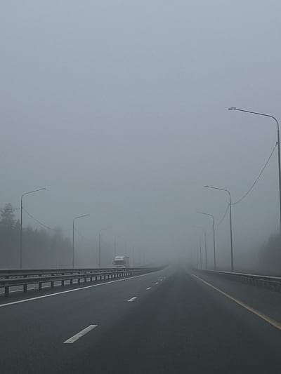 The image depicts a straight, asphalt highway shrouded in dense fog. The road has multiple lanes and lane markings. Guardrails are present on either side of the highway. Streetlights line both sides of the road, disappearing into the fog. A semi-truck is faintly visible in the distance on the highway. Trees are vaguely discernible on the left side of the road. The sky is overcast and blends seamlessly with the fog, creating a flat, gray expanse. There are no visible people. The scene appears to be in Сапуголи, Russia, based on the location provided.