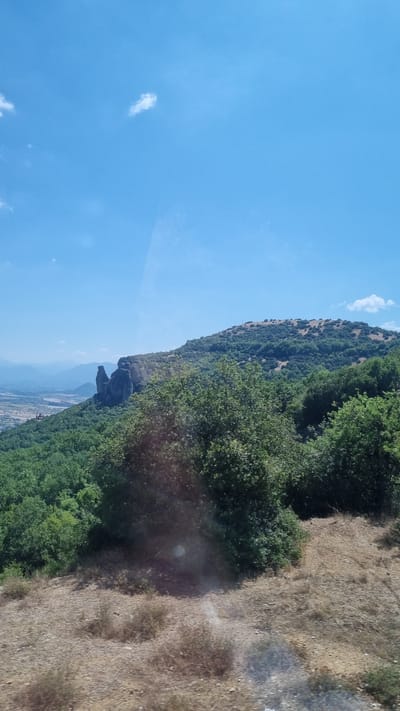 The image shows a view of the Meteora rock formations and the surrounding landscape near Kalambaka, Greece. In the foreground, there is dry, rough terrain with sparse vegetation. A dense forest of green trees occupies the middle ground, with a prominent rock formation, characteristic of Meteora, rising from the trees on the left side of the frame. The rock formation consists of several tall, weathered stone pillars. Beyond the forest and rocks, a valley with scattered buildings and further mountain ranges are visible in the distance. The sky above is clear blue with a few wispy white clouds. The overall impression is a natural landscape with a distinct geological feature.