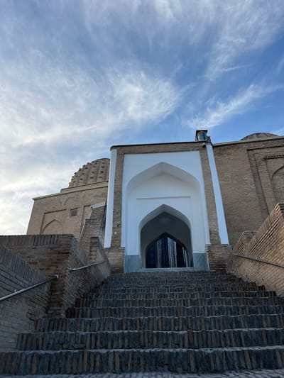 This image depicts an architectural scene featuring a brick building with a large arched entrance and a series of outdoor brick staircases. The building's facade is constructed from light brown bricks, with a prominent white arched entryway. Above the arch, there's a section of dark brickwork with decorative patterned detailing. To the right of the entrance, extensive brick stairs ascend, with metal railings providing support.
The scene is set against a backdrop of a bright blue sky with wispy white clouds, suggesting a clear day. The lighting indicates it might be late afternoon or early morning, as the light casts shadows and highlights the textures of the bricks.
The style of architecture, particularly the brickwork and the arched entryway, hints at a historical or traditional structure, consistent with the location context of Samarkand, Uzbekistan, known for its rich Islamic architecture. There are no people visible in the image. The focus is solely on the architectural elements and the sky. The overall impression is one of grandeur and historical significance.