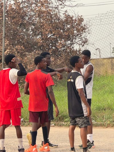 The image captures a group of six young males outdoors, appearing to be engaged in a sporting activity or warm-up. They are all standing and looking upwards, some with their arms slightly raised or positioned near their heads, suggesting they might be stretching, watching something above them, or mimicking a specific movement.
The individuals are dressed in athletic wear. In the foreground, one person wears a red short-sleeved shirt with a white design on the left shoulder and red shorts, with black sleeves or bands on their forearms and dark shoes with white socks. Next to him, another person is in a red short-sleeved top and maroon shorts, wearing black socks and bright orange athletic shoes. Behind them, another individual wears a dark short-sleeved top and dark shorts. Further back, a person is seen in a black sleeveless jersey with white trim and a white undershirt, paired with camouflage-patterned shorts, white socks, and dark athletic shoes. Part of the text "THIS W" is visible on his black jersey. Behind him, another person wears a white sleeveless jersey with black side stripes and dark shorts, dark socks, and dark athletic shoes. A sixth person is partially visible behind the group.
The setting is an outdoor area during the daytime. The ground beneath them appears to be a mix of dirt or a light-colored paved path, transitioning into green grass or low vegetation in the middle ground. A chain-link fence stretches across the background, and behind it, there are trees with numerous small, brown, dry-looking leaves, contrasting with the green grass. The sky above is bright, suggesting an overcast day or simply bright daylight. The overall impression is that of a practice session or casual gathering in an open, possibly semi-urban or rural environment.