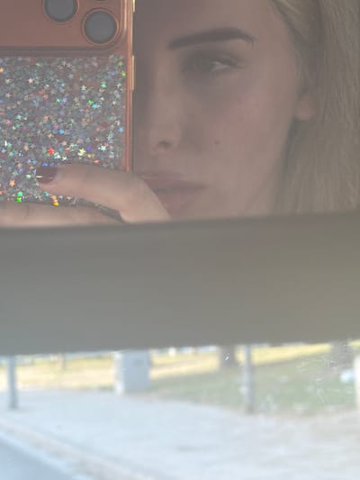 The image is a selfie taken by a woman using a smartphone. The phone, which has a rose gold colored case with a glittery star pattern, is held by the woman's hand, revealing a fingernail painted with dark red polish. She is looking into a reflective surface, possibly a mirror, as her face is partially visible. Her right eye, eyebrow, and part of her hair are visible. The background of the reflection shows an outdoor scene, possibly a street or park, with blurred trees and pavement. The setting is likely a public space in the city of Alicante, Spain, given the presence of vehicles or structures that are indistinctly visible in the blurred background. The overall image is slightly out of focus and has a hazy quality.