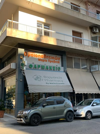 This image captures a street scene in Marousi, Greece, featuring the facade of a multi-story building that houses a pharmacy on its ground floor. The setting appears to be a typical urban environment during daylight hours, indicated by the warm sunlight casting shadows and illuminating the building's light-colored exterior.
The main subject is a pharmacy, identified by prominent signage in both Greek and English. Above the entrance, a large sign features "Ομοιοπαθητικών Μαρία Προβατά" (Homeopathic Maria Provata) in orange lettering, and "ΦΑΡΜΑΚΕΙΟ" (PHARMACY) in green lettering. A lighter-colored retractable awning extends over the storefront, also bearing text: "Φαρμακεία Pharmacy By Maria Provata." A smaller sign, partially obscured, shows "ΜΟΝΤΙΚΑ" (MONTICA). The pharmacy has a wide glass storefront displaying various products inside.
Above the pharmacy, the building extends upwards with several floors, appearing to be residential. These upper floors feature balconies with horizontal metal railings and windows, some with blinds. An outdoor air conditioning unit is visible on one of the upper balconies. Bare tree branches are partially visible on the far left, suggesting nearby foliage.
On the street in front of the pharmacy, two cars are parked parallel to the curb. The vehicle closest to the viewer is a grey SUV, possibly a Suzuki Ignis, with black alloy wheels and a partially visible license plate reading "YIT-2173". Behind it, a silver hatchback, possibly a VW Polo, is also parked. The overall impression is a quiet, sunny day on a commercial and residential street in Marousi.