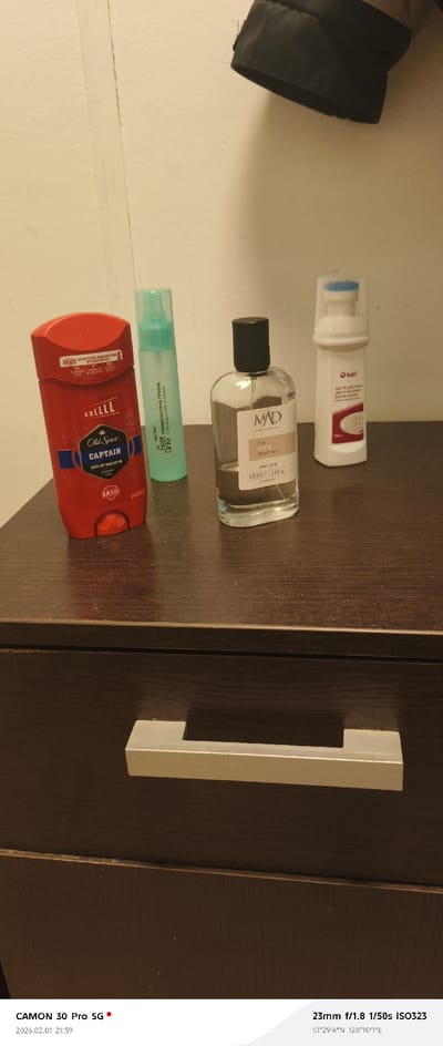 An overhead-angled photograph captures four personal care items arranged on a dusty, dark brown laminate surface, likely a dresser top, in what appears to be a residential unit within a Tecnimont camp, Russia. The specific location is indicated by coordinates 51°29'4"N 128°10'1"E, placing it near Svobodny, Amur Oblast.

From left to right, the items are: a red Old Spice Captain XXL deodorant stick; a translucent light blue-green spray bottle labeled "Feet Only" and "ПРОТИВГРИБКОВА СПРЕЙ" (antifungal spray); a clear glass bottle of "MAD PARFUMELA FOR MEN SELECTIVE" cologne, approximately two-thirds full; and a white iKari branded bottle with a blue and white pump dispenser under a clear cap, identified by Cyrillic text as a cleaning foam for white shoes (100ml e).

The background features a light-colored, possibly off-white, wall with a visible vertical seam or imperfection. A dark-colored jacket or garment is partially visible hanging in the upper right quadrant. Below the surface holding the items, a dark brown drawer front with a long, rectangular silver-colored metal handle is visible. No people are present.