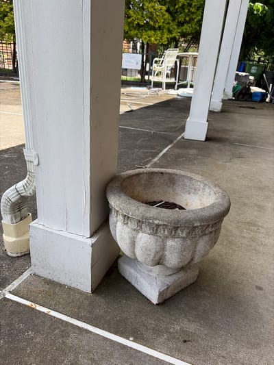 A partially obscured, medium shot depicts an exterior location in Edison, United States. The scene prominently features a weathered, gray stone or concrete urn resting on a painted white wooden beam. The urn sits slightly askew, revealing some internal debris or dried plant matter. The beam is adjacent to a white, rectangular column, likely part of a porch or patio structure. A white downspout runs along the bottom of the beam. The concrete surface appears well-worn and gray, with subtle surface variations. In the blurred background, there are glimpses of greenery, white patio furniture, and a green trash container, suggesting a residential or commercial outdoor space.