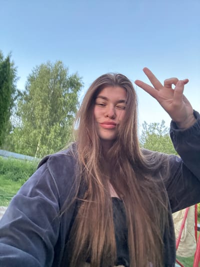 A young woman with long, brown hair is taking a selfie outdoors. She is wearing a dark-colored jacket over a black top. Her right hand is raised, and she is holding up two fingers in a peace sign. She is winking and has a slight smile. The background is blurred, showing trees and a clear blue sky. The scene appears to be a residential area in the city of Meshchera, Russia, possibly a backyard or park.
