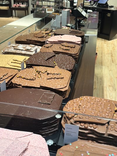 The image depicts a display case filled with various types of chocolate slabs, likely in a confectionery or chocolate shop. The chocolates vary in color and texture, some featuring nuts and other toppings. A small label, partly visible, identifies one type as "Macadamia." The location is suggested to be in Spreitenbach, Switzerland. The scene is viewed through the glass of a display case, with reflections on the surface. The background includes a blurred view of a store interior.