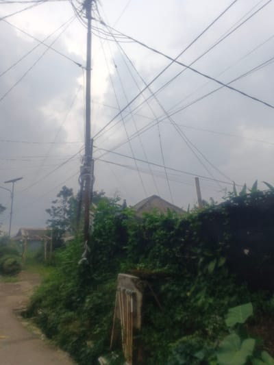 The image depicts an outdoor scene in Morrumbala, Mozambique, under an overcast sky. A prominent, dark utility pole stands vertically in the center-left, laden with numerous intertwined electrical or communication wires that crisscross the upper frame. Attached to this pole are various electrical components, including what appears to be a transformer. To the left, an unpaved path or road extends into the background, where another utility pole, topped with a solar panel, and a rustic, open-sided structure with a light roof are partially visible amidst green trees. The foreground and midground on the right are dominated by dense, vibrant green foliage, including shrubs and vines, obscuring a lower wall or bank. Partially visible within this vegetation in the lower right is a concrete structure, possibly a gatepost, with vertical metallic elements. Further into the background on the right, behind the foliage, the conical or pyramidal thatched roof of a building is visible. No individuals are present, and no discernible actions or interactions are occurring. The environment suggests a semi-rural or peri-urban setting with basic infrastructure and extensive natural growth.