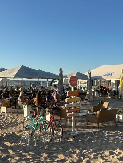 This media file captures a lively outdoor cafe scene on a sandy beach, likely in Jūrmala, Latvia, under a clear blue sky. The time of day appears to be late afternoon or early evening, with the golden sunlight casting long shadows across the sand and wooden boardwalk. Several people are seated at tables, enjoying drinks and conversation, suggesting a relaxed social gathering.
Prominent objects in the scene include tables and chairs, some of which are made of woven wicker, characteristic of beachside establishments. Umbrellas provide shade for the seating areas. A bicycle rack holds a couple of bicycles, one a bright turquoise and another red, hinting at the recreational nature of the location.
Several signs with directional arrows add to the festive atmosphere. One sign reads "SUMMERTIME WITH APEROL SPRITZ," promoting a refreshing drink. Another sign cluster features phrases like "NOTHING OUTSHINES A SPRITZ SMILE," "mix with friends," "ORANGE VIBE," "Better Together," and "APERITIVO TIME," all conveying a sense of enjoyment and socializing. The overall impression is one of a vibrant and appealing spot for relaxation and enjoying the beach.