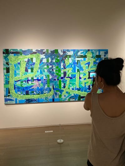 A person, viewed from behind, stands in a well-lit art gallery, holding a mobile phone to take a picture of a large, abstract painting. The painting is composed of various shades of blue and green, with thick, bright green brushstrokes forming what appear to be stylized characters. The person is wearing a light-colored tank top and large hoop earrings, with their dark hair pulled up in a bun. The gallery floor is made of polished wood. A small white sign with some text on it is visible on the floor near the painting. The painting is hung on a white wall. The scene takes place in the Jinniu District, China.