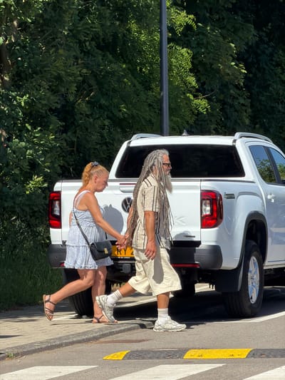 Two people are walking along a curb, next to a white Volkswagen Amarok vehicle, license plate RY2. The woman is on the side closest to the vehicle, wearing a blue and white dress, dark sandals, and a black purse. The man is walking slightly ahead of her, wearing light-colored clothing, white socks, white sneakers, and has long dreadlocks. Behind them, there is a lush green area. The curb is marked with yellow and black stripes. The location is Fentange, Luxembourg, as inscribed on the license plate.