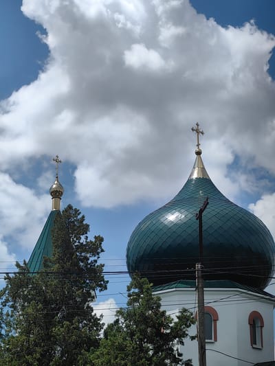 The image shows the domes of an Orthodox church in the city of Zuya, Ukraine, against a backdrop of a partly cloudy sky. A larger, emerald green dome with a golden cross is in the foreground on the right, and a smaller, similar dome is visible on the left, also topped with a golden cross. The church structure is white with arched windows featuring reddish trim. Green trees obscure the lower portions of the building and extend upwards towards the domes. Electrical wires and a utility pole are present in the mid-ground, intersecting the view of the church and trees. The scene is captured from a low-angle perspective, emphasizing the scale of the church against the sky.