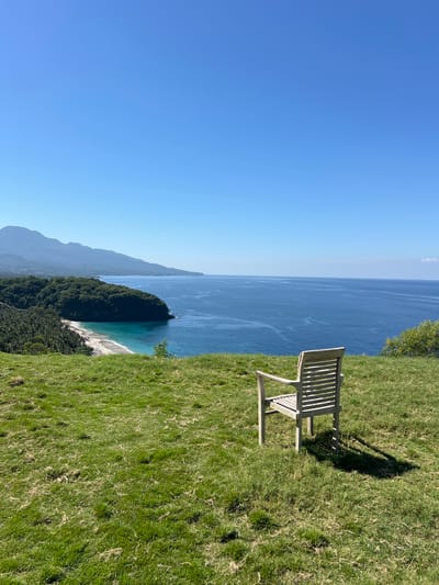 The image depicts a scenic overlook in Karangasem, Indonesia. In the foreground, a weathered wooden bench sits on a grassy knoll. The bench faces the ocean, which stretches out to the horizon under a clear blue sky. In the middle ground, a crescent-shaped beach is visible, with turquoise water lapping at the shore. Lush green trees and vegetation cover the cliffs surrounding the beach. In the background, a large mountain range rises majestically, its peaks partially shrouded in haze. The overall scene is one of tranquility and natural beauty.