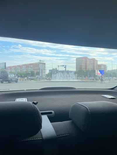 The image shows a view from inside a car, looking out the rear window at an urban scene. The window has horizontal defroster lines. Outside, there are buildings, a street with cars, and a crowd of people. The sky is blue with white clouds, suggesting daytime. A fountain is visible in the distance, and some of the buildings appear to be residential blocks. On the right side of the image, car seats are visible, including a grey seatbelt. There is some text on a label attached to the car interior, but it is not legible. The overall impression is of a sunny day in a city, possibly during an outdoor event or gathering. The image appears to be taken in Pavlodar, Kazakhstan.