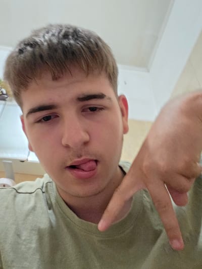 The image is a close-up selfie of a young person with short, light brown hair and a light olive-green t-shirt. The person's tongue is sticking out to the right, and their right hand is held up in front of their chest, with the index and middle fingers extended to form a "V" shape. The background appears to be an indoor setting, possibly a home, with white walls and a ceiling visible. There is no visible indication of the city of Aleksandrovo, Bulgaria.