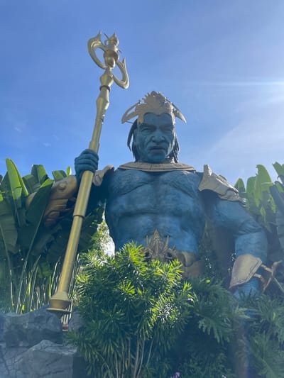 A low-angle, eye-level shot captures a colossal blue statue of a mythical warrior, likely of oceanic or aquatic origin, holding a golden trident. The statue is adorned with ornate gold armor on its shoulders and chest. It stands amidst lush green foliage and dark, textured rocks, possibly in a garden or themed attraction. The sky above is a clear blue with the sun creating a lens flare effect on the right side. The statue's facial features are stern and imposing, with dark, braided hair and a golden headdress. The image provides a detailed, realistic portrayal of this impressive monument.