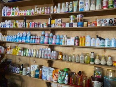 The image depicts a retail shelf in what appears to be a small convenience store or market stall, likely located in Ta', Indonesia. The shelves are stocked with a wide assortment of personal care products, household items, and some food items.

The top shelf displays various brands of body wash, lotion, and other liquid personal care products in plastic bottles and tubes. Brands like Marina, Citra, and Herbalist are visible.

The second shelf from the top is filled with more personal care items, including shampoos, conditioners, and styling products. There are also some boxed items, possibly hair dye or other beauty treatments, with brands like Miranda prominently displayed. Small packaged food items are also present, such as instant noodles in cups.

The third shelf contains a mix of baby powder, body lotion, and other hygiene products. Several brands of baby powder, such as "Cussons baby," are noticeable in their distinctive packaging. Small bottles and jars of lotions and creams are also arranged here.

The bottom shelf, which is partially visible, shows more personal care products, including mouthwash (Listerine), body wash, and fabric softener pouches. There are also jars containing small, light-colored, spherical objects, possibly decorative or consumable items. Some larger plastic containers and a jar of candy are also visible on this shelf.

The environment is a wooden shelving unit, suggesting a simple retail setup. The lighting is typical of an indoor setting, possibly with overhead lights or natural light from an unseen source. The overall impression is one of a well-stocked, though potentially cluttered, retail space. No people are visible in the image.