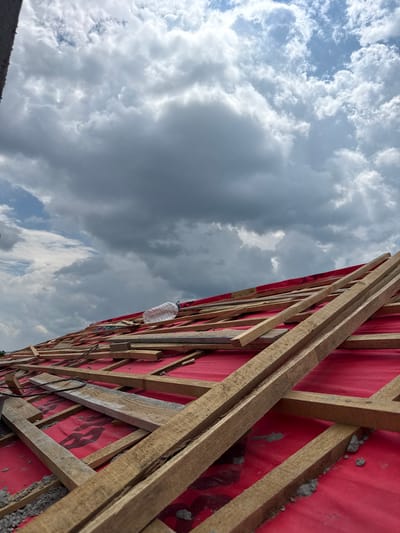 The image shows an unfinished roof covered with red protective sheeting and wooden battens. Several pieces of wood are scattered across the roof, and a plastic water bottle rests on top of the battens in the upper right quadrant of the image. The sky above is filled with dramatic, dark clouds, suggesting an impending storm. The location is identified as Ovoshtnik, Bulgaria.