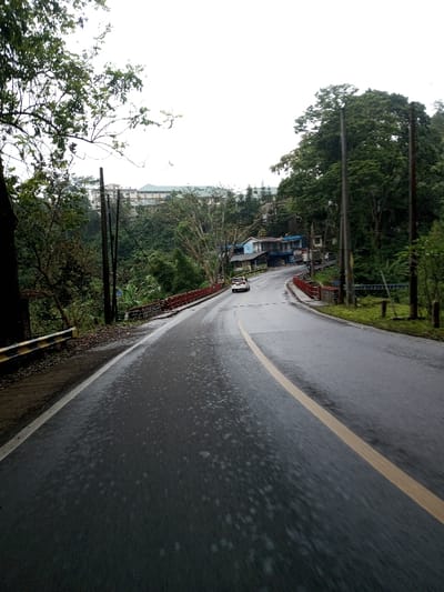 The image depicts a wet, winding asphalt road in YonPhoogla, Bhutan, on an overcast day. The road, marked with a yellow center line and a white shoulder line on the left, extends from the foreground, curving gently to the left before ascending. Its surface shows numerous damp patches, indicative of recent rain.
On the immediate left, a yellow and black striped guardrail runs alongside dense green foliage. Further along the road on the left, multiple dark utility poles stand amidst trees. A bridge with red railings crosses a dip in the terrain ahead.
On the right side of the road, near the bridge, a cluster of small, multi-story buildings with blue and light-colored facades is visible. A white sedan is present on the road, moving away from the viewer, past these buildings. More utility poles are present on the right, interspersed with abundant green trees and vegetation.
In the background, positioned atop a forested hill, a large, multi-story building or complex with a light facade and dark roof is partially visible through the trees. The sky is uniformly bright and overcast. No individuals are discernible.