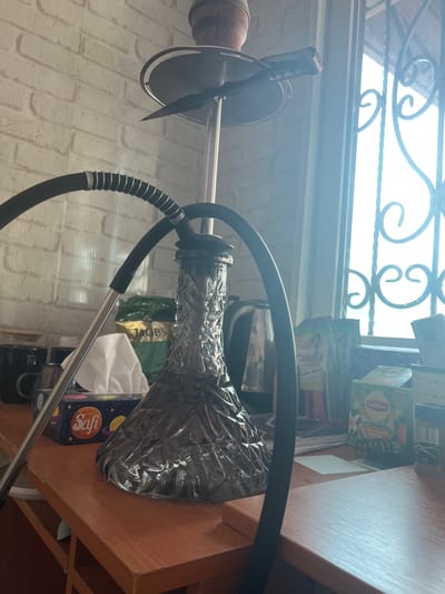 The image shows a hookah setup on a wooden shelf next to a window with decorative metal bars. The hookah has a dark, textured glass base and a silver stem. A black hose with a coiled metal grip is attached to the hookah. In the background, several boxes of tea, including Lipton and Jacobs, are visible. A box of Safi tissues with images of planets is also on the shelf. The scene appears to be indoors, with natural light coming from the window, suggesting it could be daytime. The white brick-patterned wall in the background provides further setting detail. There is no visible activity or people in the image. The overall impression is one of a domestic setting where a hookah is prepared or has been used.