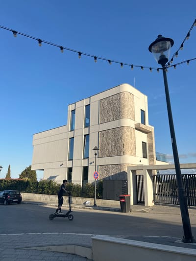 This image captures an outdoor scene in Chernomorets, Bulgaria, under a clear blue sky, suggesting it is daytime and the weather is fair. The central focus is a modern, multi-story building with a distinctive architectural design, featuring curved lines and a combination of light-colored stone and smooth, cream-colored facade elements. Large, rectangular windows punctuate the building's exterior.
In the foreground, a person is seen riding a black electric scooter on a paved road. The rider is wearing dark clothing and appears to be casually moving along the street. Adjacent to the road is a sidewalk constructed with light-colored paving stones. A black trash bin with red lettering is visible near the building entrance.
To the left, a lamppost with a decorative lantern is prominent, adorned with a string of small, exposed light bulbs that add an aesthetic touch to the scene, even though it is daytime. A traffic sign, a red circle with a diagonal red bar, indicating a prohibition, is also visible on a pole.
In the background, behind the building, a black car is parked on the street, and further beyond, lush green trees and vegetation are visible. The overall impression is one of a contemporary urban or semi-urban environment on a bright, sunny day.