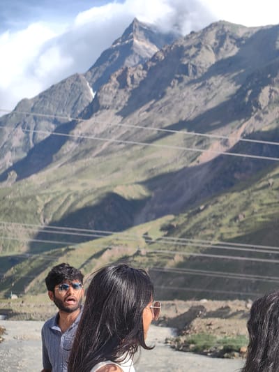 This photograph, taken in the city of Yamling, India, captures a scenic mountain landscape under a partly cloudy sky. In the foreground, a man wearing sunglasses and a blue striped shirt is looking to his left with his mouth open. To his right, the back of a woman's head with long dark hair is visible. The background is dominated by a vast mountain range with steep, rocky slopes covered in patches of green vegetation. Several power lines are strung across the middle ground, traversing the mountain face. The overall scene suggests an outdoor adventure or a scenic viewpoint in a mountainous region.