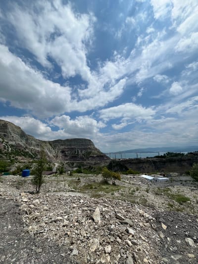 The image shows a landscape with a rocky, mountainous terrain and a cloudy sky, taken near Chirkey, Russia. A slope of rocky material dominates the foreground. In the background, a tall, rocky mountain range is visible under a sky filled with cumulus clouds. A bridge is faintly visible crossing the water in the distance. Power lines stretch across the sky. Vegetation dots the landscape. A blue barrel is near the base of the mountain. In the far background, a settlement or industrial complex is present with long, white buildings.