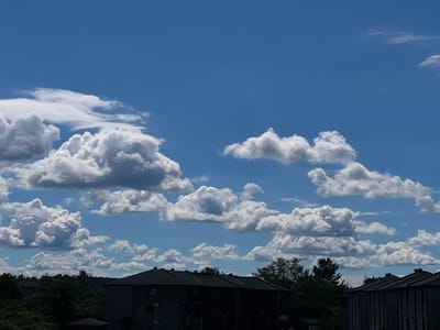 The image shows a view of apartment buildings under a bright blue sky with numerous white cumulus clouds. The buildings are dark-colored with peaked roofs and appear to be residential units. Trees and foliage are visible in the foreground and between the buildings. The scene is set in the city of Gatineau, Canada, as indicated by the surrounding architecture and vegetation, which are consistent with the region. The time of day appears to be daytime, likely mid-morning or afternoon, given the bright lighting and clear sky. There are no people or animals visible in the frame.