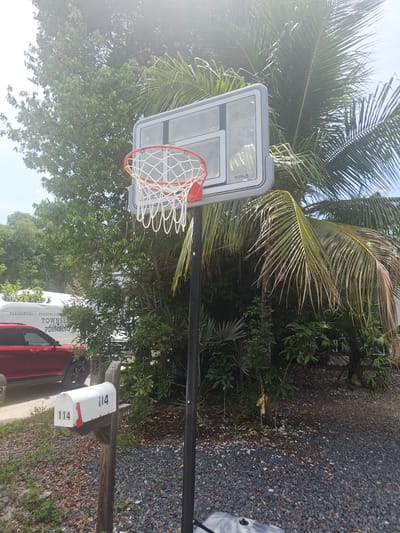 In Islamorada, Florida, a basketball hoop with a white and red rim stands next to a mailbox. The mailbox, numbered 114, is made of white metal and mounted on a wooden post. A red SUV is parked behind the mailbox. Lush green trees and palm trees surround the scene, with a bright, hazy sky overhead. The ground is covered in gravel and fallen leaves.