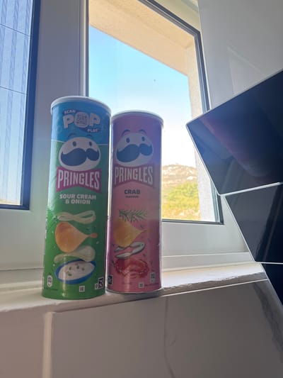 The image shows two Pringles cans, one Sour Cream & Onion and one Crab flavor, positioned on a windowsill with a view of the outdoors. The Sour Cream & Onion can is green and features an illustration of chips with sour cream and chives. The Crab flavor can is pink and depicts crab, rosemary, and a crab claw. The windowsill itself appears to be part of a modern interior, with a reflective surface below. Outside the window, a bright blue sky is visible, along with a hint of vegetation and possibly distant buildings, suggesting a sunny day. The location is identified as Sustaš, Montenegro. There are no people or animals present in the image. The lighting suggests it is daytime, with natural light coming from the window.