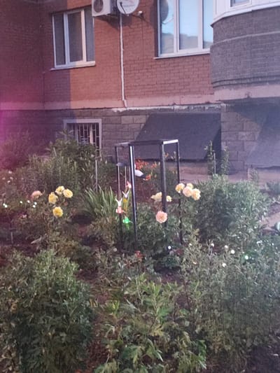 The image depicts a garden area in front of a brick building, possibly in Shchyolkovo, Russia. The building has a red brick facade with a white-framed window and an air conditioning unit visible near the top. Below this, there is a smaller window with a security grate. The garden itself is lush with various plants and flowers, including several yellow and pink roses. Small, colorful decorative ornaments, possibly paper cranes, are hanging from a metal structure in the garden, along with some string lights that add a festive touch. A bird feeder is also mounted on a pole. The lighting in the image suggests it is either dusk or early evening, with some artificial lights illuminating the scene. The overall mood is peaceful and decorative, indicating a well-maintained outdoor space. There is no visible text in the image.