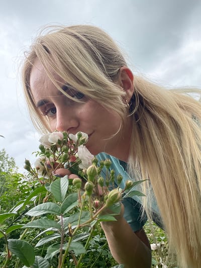 A young woman with long blonde hair is in a garden, smelling a small bouquet of pink and white roses. She is wearing a teal-colored top. The background is filled with greenery and a cloudy sky. The scene is likely in Yasnoe, Russia, a town known for its natural beauty and gardens.