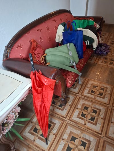 The image depicts a cluttered interior scene. A red, ornately designed sofa is piled with various items including clothing, bags (one green and one blue), and an umbrella. The sofa has a floral pattern upholstery. To the side of the sofa is a white table with flowers in a vase on top. The floor is tiled with a complex geometric pattern of light and dark brown. The location and the city of Krumovgrad, Bulgaria cannot be determined from the image.