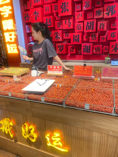 A woman wearing a gray t-shirt stands in front of a stall selling red beads in a market in Hangzhou Shi, China. The stall has rows of clear glass containers filled with the red beads, and there is a sign written in Chinese above each container. Behind the woman is a wall of red square tiles with Chinese characters written on them. The woman is holding a small fan in her right hand and pointing at the red beads with her left hand. There is a white rectangular box sitting on the counter, and a sign above it that is also written in Chinese. The stall is located on the ground floor of the market, and there are other stalls nearby. The market is brightly lit, and the walls are a light beige color. There are a few people visible in the background, but they are blurry. The stall is a popular spot in the market, as there are several customers browsing the red beads. The red beads are displayed in a neat and organized manner, and the stall appears clean and inviting.