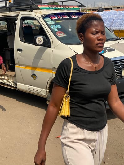 An adult female is captured in a close-up, walking outdoors in Ablekuma, Ghana, with her eyes closed and a neutral expression. She has short, reddish-brown hair and wears a black, short-sleeved scoop-neck top, light-colored (possibly off-white or light grey) casual trousers with a white drawstring, a gold-colored necklace with a small pendant, and a small earring in her left ear. A yellow, crocodile-textured handbag with a gold-colored chain strap is carried over her right shoulder.

Behind her is a light-colored (cream/off-white) passenger van, likely a commercial minibus or "trotro," which occupies a significant portion of the background. The van features a roof rack, a Ghanaian flag-themed stripe above the windshield with "REMEMBER" written on it, and "BIG BOSS" prominently displayed on the windshield visor. Multiple stickers, including one for the "Driver and Motor Transport Union" and a Real Madrid C.F. logo on the right-side mirror, adorn the front. A yellow stripe runs along the lower side panel, which also bears text "Spirit mind" and "Produced by TOYOTA SPORT." The side door is open, and a hand in a red sleeve is visible within the opening. The van shows signs of wear, including minor dents and dirt. In the background, beyond the van, blurred structures and stacks of what appear to be bottled water are visible under a hazy sky. The ground is an unpaved or dirt-covered surface.