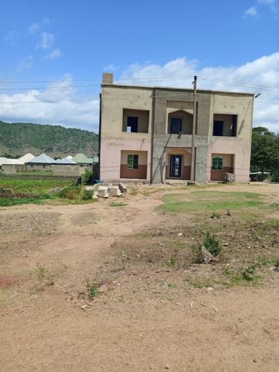 The image depicts a two-story building under construction in Barakin Babele, Nigeria. The building has a beige and light pink exterior with dark gray accents around the windows and entrance. Several windows are visible, some are open voids and others have dark blue doors or frames. A prominent dark blue door is at the center of the ground floor, accessed by a short set of concrete steps. The surrounding environment is dry and dusty with patches of green grass and vegetation. In the background, a lush green, hilly landscape with some smaller structures with metal roofs is visible under a bright blue sky with scattered white clouds. Electrical wires are strung across the upper portion of the image. The ground in the foreground is a dirt road with scattered rocks and debris.
