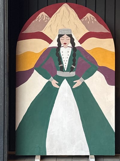 This media file is a photograph of a painting, likely a decorative piece, mounted on a dark surface. The painting depicts a stylized figure of a woman in traditional attire, lying down with her eyes closed and a peaceful expression. She has long, dark braided hair adorned with what appear to be small, sparkly embellishments and a fur-trimmed hat. Her clothing is colorful, featuring a green dress with purple accents and a white underskirt.

The background of the painting is abstract, composed of curved, layered bands of color in shades of red, yellow, and cream, which evoke a sense of a mountainous landscape or a sunset. The overall style is folk art, characterized by bold colors and simplified forms.

The painting is oriented horizontally, and its curved left edge suggests it might be part of a larger installation or a unique architectural element. The photograph is taken from a slightly elevated, frontal perspective, with a dark frame or border surrounding the artwork. The lighting appears to be natural, casting soft shadows and highlighting the texture of the paint. No text is visible in the image. The setting is likely indoors, given the controlled lighting and the visible mounting mechanism. The artwork is situated in Voronezh, Russia.
