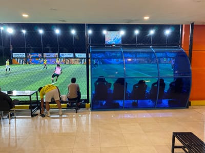 The image depicts an indoor artificial turf football field at night in Banda Aceh, Indonesia, illuminated by bright overhead lights. The field is bordered by a net fence. On the field, several young men are playing football, dressed in various team uniforms. One player in a yellow jersey and white shorts is on his hands and knees near the sideline, appearing to be picking something up. Another player in a pink jersey and black shorts is running. Spectators are seated on a bench to the right of the field, behind a blue-tinted transparent enclosure that houses additional seating. The floor in the foreground is tiled with a reflective surface. Advertisements and graphics are visible on the fence surrounding the field.