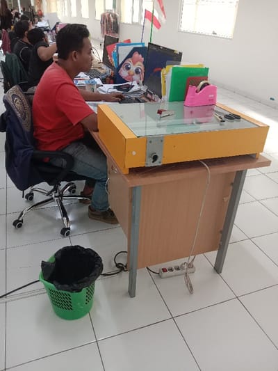 The image shows a man sitting at a desk in an office. The man is wearing a red shirt and blue jeans, and he is using a computer with a yellow-and-glass top. He is positioned in a cubicle, which is lined with white tiles. The cubicle is in a larger office, which appears to have large windows with a view of another building. There is a black trash can with a green liner to the man's left. The image was likely taken at an office in the Kecamatan Ngaliyan district of Semarang, Indonesia, based on the provided coordinates.  A power strip is visible on the floor underneath the desk. Several colorful binders are visible on a shelf behind the man. A red-and-pink tape dispenser sits atop a stack of orange and green binders. A red object is visible on the surface of the desk, in front of the man.  The image is taken from a slightly elevated angle, and the focus is on the man and his desk.  The image appears to have been taken in natural light.  There is no visible interaction between the man and any other people.