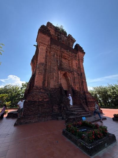 In the city of Tuy Hoa Ward, Vietnam, a tall, ancient brick tower stands under a clear blue sky with sparse white clouds. The tower, constructed of reddish-brown bricks, features a stepped base and a central arched entrance. Vegetation grows atop and around the structure. Several people are present: a man in a white shirt and khaki shorts leans against the tower's base on the left. To his right, a woman in a pink scarf and a black outfit walks away from the tower. In the center, on the steps leading up to the entrance, a person in white clothing ascends, followed by another person in a colorful shirt and jeans who appears to be taking a photo with a mobile device. A planter box filled with vibrant red flowers sits in the foreground on the right, surrounded by a tiled courtyard. Trees and foliage form the backdrop of the scene.