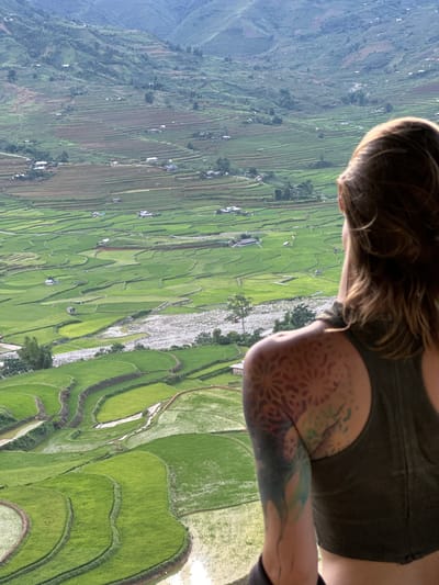 An individual is depicted from a rear-over-the-shoulder perspective, observing a vast terraced landscape in Tú Lệ, Vietnam. The person has medium-length brown hair and wears a dark green sleeveless top. A patterned tattoo, featuring a geometric, mandala-like design in reddish-orange tones, is visible on their left shoulder. Below it, on the upper left arm, is another tattoo displaying abstract forms with blue, green, and yellow hues. The expansive valley below is characterized by numerous vibrant green rice terraces, some partially flooded, conforming to the undulating terrain. A light grey, rocky riverbed, containing visible water, courses through the midground. Scattered throughout the rice fields and on the lower slopes of the hills are multiple small, simple structures, identified as dwellings. The distant background consists of densely vegetated mountains beneath a hazy, light blue sky.