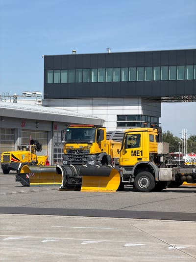 The image shows a bright, sunny day with a clear blue sky. The scene is an outdoor area, possibly an airport or industrial facility, given the presence of large vehicles and a modern, multi-story building with numerous windows.
The primary subjects are two yellow trucks equipped with snowplows, suggesting the image was taken during a period when snow removal equipment is active, or for storage. The trucks are parked side-by-side. The first truck, closer to the building, is larger and has "ME1" and "2043" written on its side. The second truck is partially obscured by the first and also features yellow snowplow attachments. The building is dark gray with horizontal bands of windows and appears to be a functional facility, possibly an office or control tower. There are some red numbers visible on the building's exterior, "37" and "38", which might indicate bay numbers or sections.
The ground is paved, with clear lane markings visible on the right side of the image, further supporting the idea of a transportation hub or large operational area. The overall atmosphere is one of industrial activity and preparedness, with no visible people. The lighting indicates it is daytime, likely midday due to the harsh shadows and bright illumination. The location is specified as Balice, Poland, which is known for its airport.