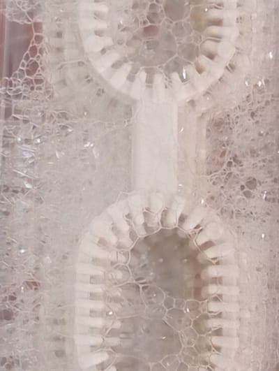 The image shows a close-up view of soap bubbles and a bubble wand. The wand appears to have two circular openings with ridged edges, designed to create multiple bubbles at once. The bubbles are a translucent white and are clustered around the wand, creating a frothy texture. Some small, bright reflections are visible within the bubbles, suggesting they are catching light. The background is out of focus and appears to be a soft, neutral color. The image does not contain any discernible elements that would indicate its location in the city of Smila, Ukraine.