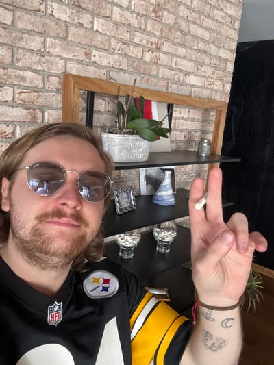 A Caucasian male with long blonde hair, a mustache, and a beard is seen taking a selfie. He is wearing round sunglasses and a Pittsburgh Steelers jersey. He holds a thin, white cylinder object, resembling a cigarette or a joint, between his fingers. His right forearm is visible, adorned with a woven bracelet and tattoos.
Behind him, there is a shelf displaying decorative items, including a potted plant labeled "Flowers & Garden," small decorative vases and containers. The shelf is positioned against a brick wall.
The image provides no information to ascertain the location in Huixquilucan de Degollado, Mexico.