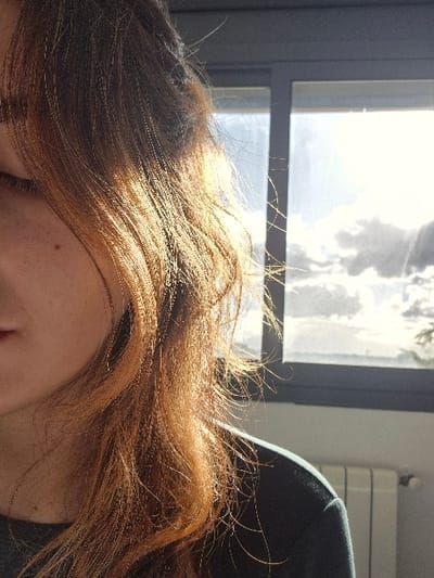 The image depicts the left profile of a person indoors in Tixeraïne, Algeria. The individual has long, wavy, reddish-brown hair, heavily illuminated by direct sunlight from a large window positioned to their right. The left eye is partially visible, appearing closed or directed downwards, with a small dark mark present on the cheek below the eye. The person is wearing a dark green long-sleeved garment. Beyond the dark-framed window, a bright sky with visible cloud formations is evident. A white, ribbed radiator is situated directly below the window on the right side of the frame.
