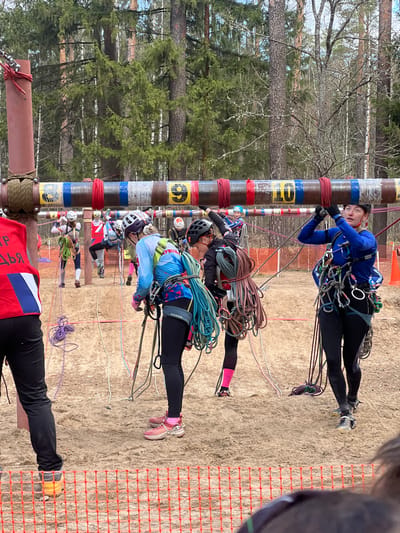 The image depicts an outdoor climbing competition in Lesnoye, Russia. Several individuals wearing helmets and climbing gear are ascending numbered poles. They are secured with ropes and harnesses. The poles are marked with alternating bands of red, white, and blue, with yellow numbers indicating their position. The background includes a wooded area and a sandy ground surface. A safety fence is visible on the right side. A person wearing a red and blue vest is visible at the bottom of the image.