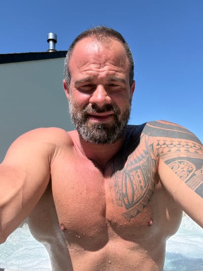 The image is a selfie of a shirtless man with a muscular build, a beard that is graying, and tribal tattoos on his left shoulder and upper arm. His skin appears to be wet, possibly from water or sweat. The sky is visible in the background, along with a white building or structure. A gray object is affixed to the building, perhaps a vent. The photo appears to have been taken outdoors in daylight. The location in the city of Marche-en-Famenne, Belgium, cannot be confirmed based on the provided image alone.