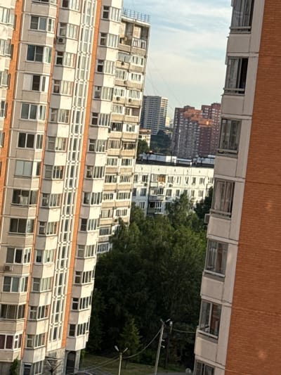 This image shows a view of several apartment buildings in the city of Kotelniki, Russia. The buildings are multi-story structures with numerous windows and balconies. The closest building on the right is made of red brick. A large number of green trees are visible between the buildings, suggesting a moderately dense urban environment with significant green spaces. The sky is partly cloudy. No people are visible in the image. The scene appears to be a daytime, outdoor view.
