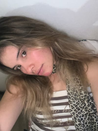 A woman with long, light brown hair is positioned at an angle in the frame. Her face is visible, and she is looking toward the camera. She is wearing a leopard-print top that transitions into a white and brown striped garment. The background includes a white wall on one side and appears to be indoors. There is no indication of the location being Sumalao, Argentina, or any other specific geographic location.