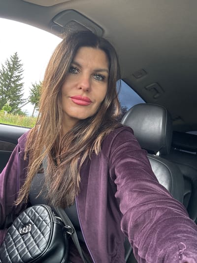 The image shows a woman sitting in the driver's seat of a car. She has long, brown, wavy hair, blue eyes, and is wearing makeup. She is wearing a purple velour tracksuit over a black top and has a black quilted handbag with silver hardware on her lap. The car appears to be parked outdoors, with trees and some greenery visible through the window. The scene suggests a casual, everyday moment. The location is identified as Kvarts, Russia.